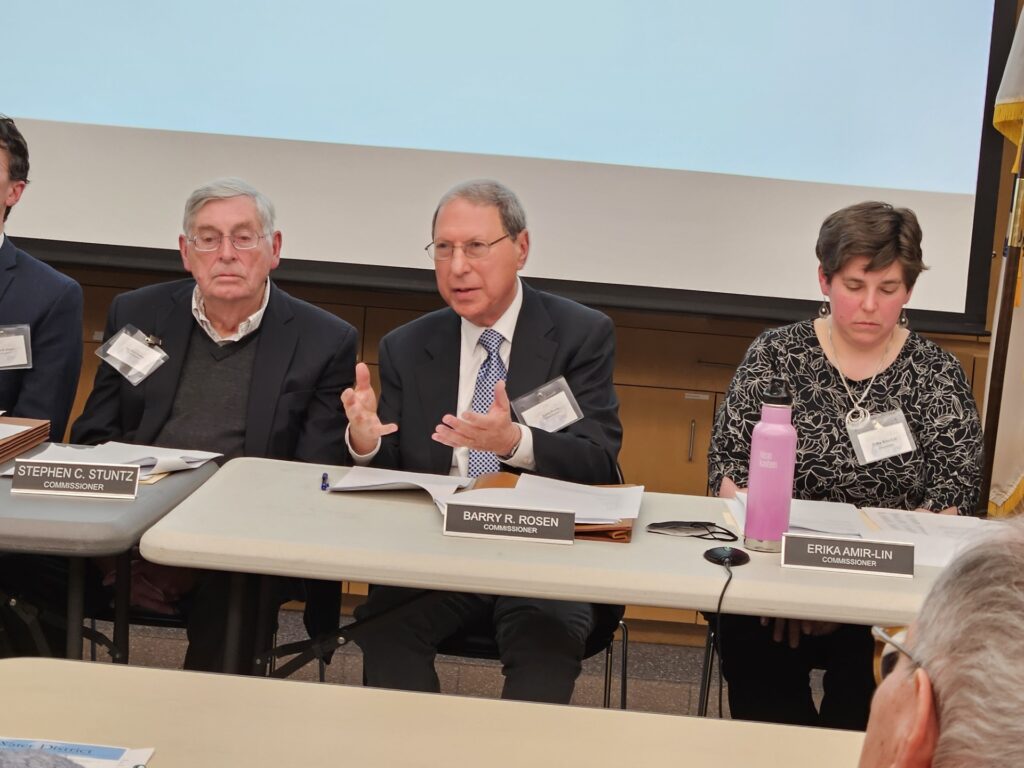 Two men and a woman sit at desks in front of a large screen. They each have nameplates on the desk that indicate their names and roles (Commissioner).