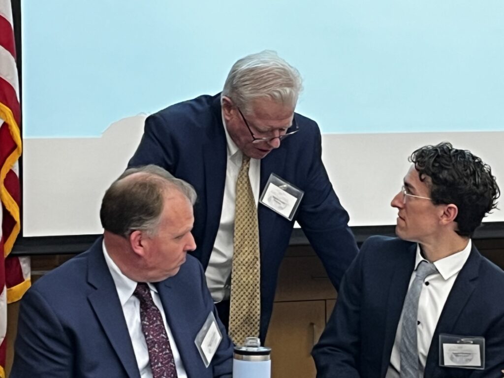 Three men in suits are at a table. The man in the middle (the moderator) is standing over the other two, who are seated and facing him.