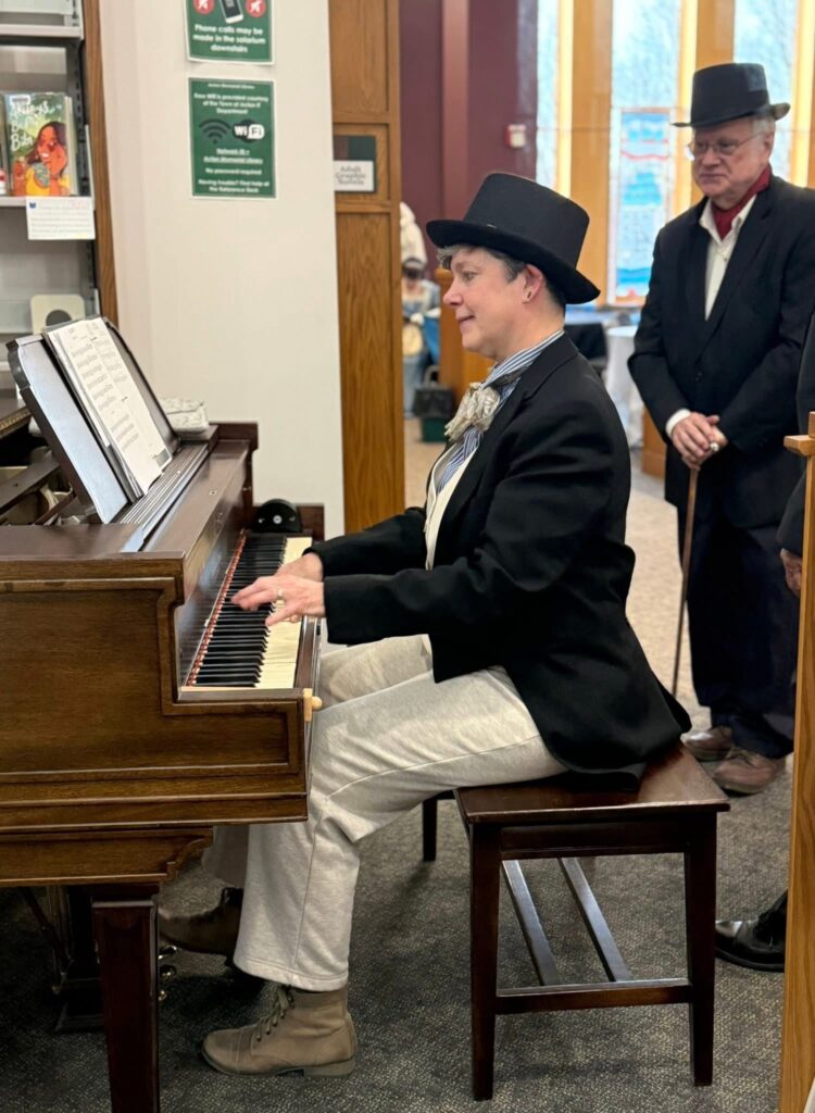 A short-haired woman wearing a top hat, striped shirt with a floppy tie and vest, and black cutaway jacket plays the piano. A man also wearing a top hat and black suit stands behind her.
