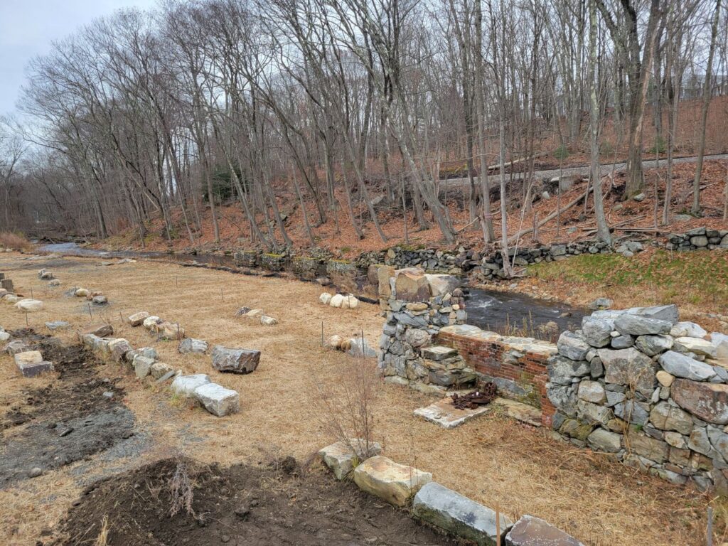 In the background, a stream runs off into the distance. In the forground, stones, and a brick and stone wall.