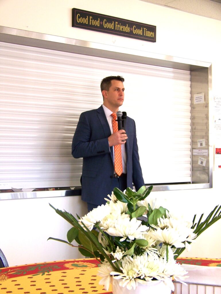 A tall man in a suit and orange polka dot tie stands in front of wall at the Senior Center. He's holding a couple of mics in his hand.