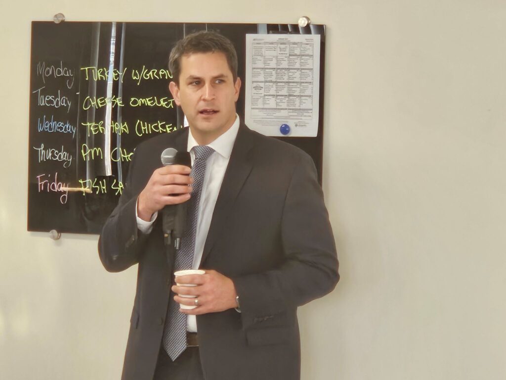 A man in a tie speaks into a pair of microphones while holding a cup of coffee.