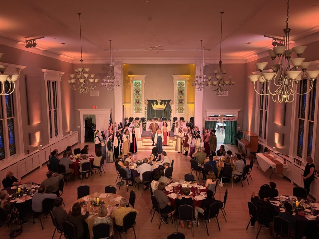 A hall with a small stage up front. on each side of the stage, singers in medieval garb stand on the steps. There are a number of round tables in the main hall, where people are listening to the Madrigals singing.