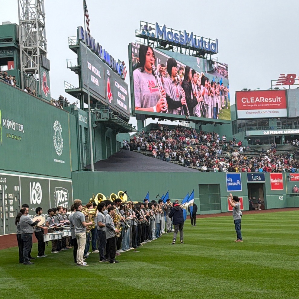 A group of musicians wearing grey t-shirts is dwarfed by the Green Monster wall at Fenway park. The Jumbotron on the right shows the musicians up close.