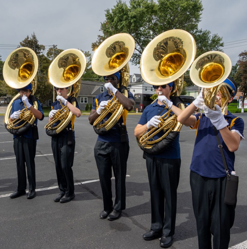 Five brass players in a casual uniform(a blue t-shirt and white gloves) get ready to march from a parking lot.