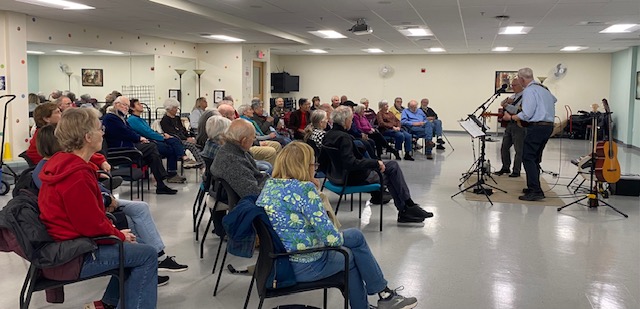 A large audience sits in a hall listening to two musicians at the front.
