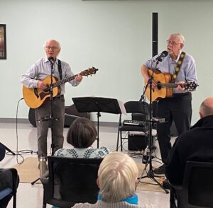 Two older men paly acoustic guitars and sing in to microphones.