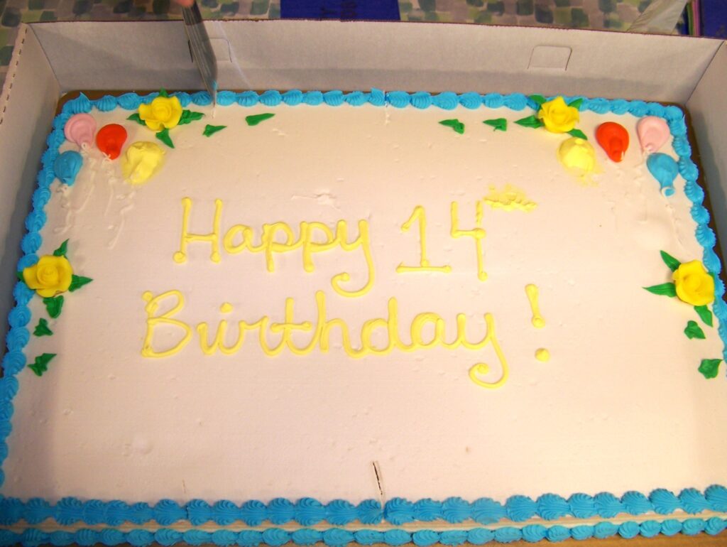 A large white-frosted cake decorated with flowers and yellow writing that says "Happy 14th Birthday!"
