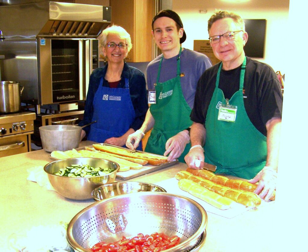 A women and two men stand in front of bowls of salad and trys of garlic bread. All theree are wearing aprons.