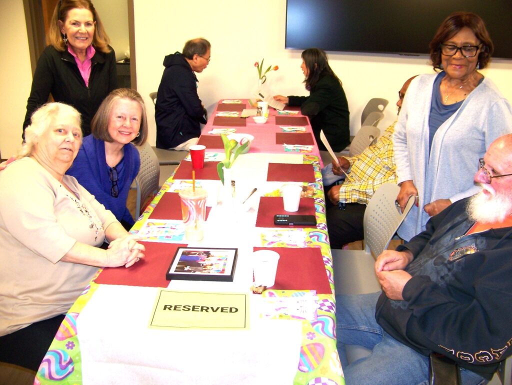Several people sit at a table that has Easter decorations. One woman stands over two seated women on the right. In the background some other guests are chatting at a table.