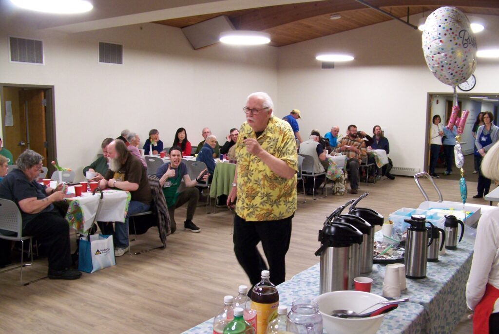 An older man wearing a bright yellow Hawaiian shirt speaks into a microphone in the middle of a hall. People are sitting at tables listening (and eating dinner).