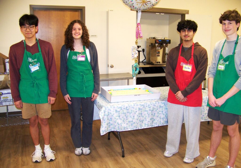 Four smiling high-schoolers stand in front of the cake wearing aprons.