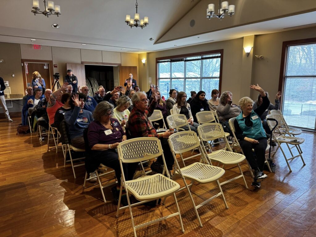 A room full of people sitting in white plastic chairs. Some people have their hands raised.