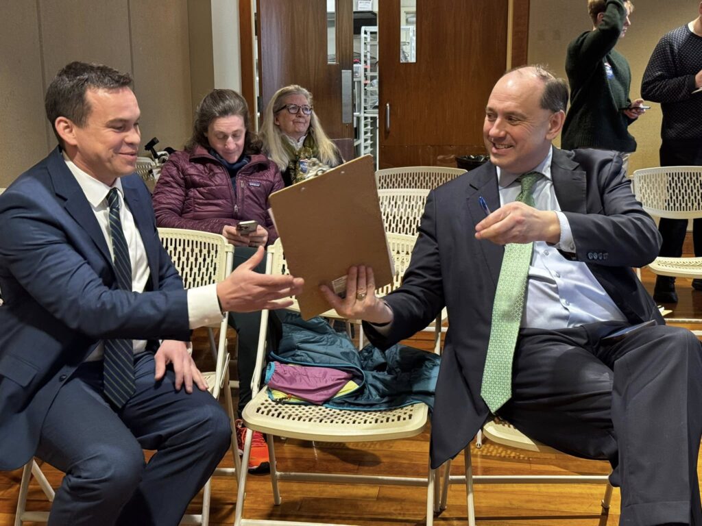 Two men in suits are sitting in a meeting room. One man is handing the other a pen and clipboard. Behind them, people are listening, chattign, and checking their phones,