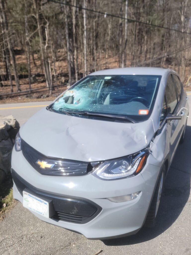A gray car with a a bunch of dents on the hood and front and a large hole in the windshield.