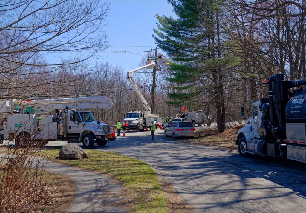 A long distance shot of several trucks parked on and near the road. Two workers are up in cherry pickers workiing on a utility pole.