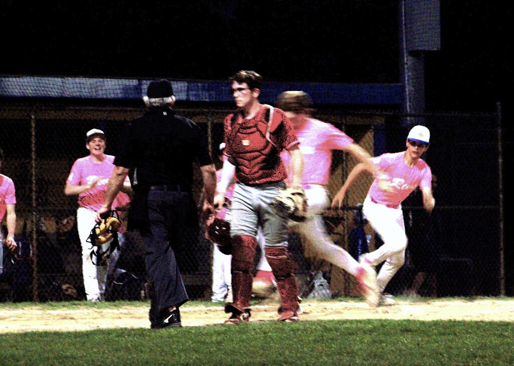 In the foreground, a catcher walks across the field. Behind him, a player is running into home plate.