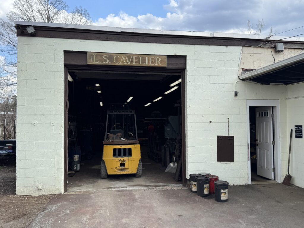 A concrete building wwwith a wooden sign that says L.S. Cavelier. A yellow forklift is inside the open garage-style door.