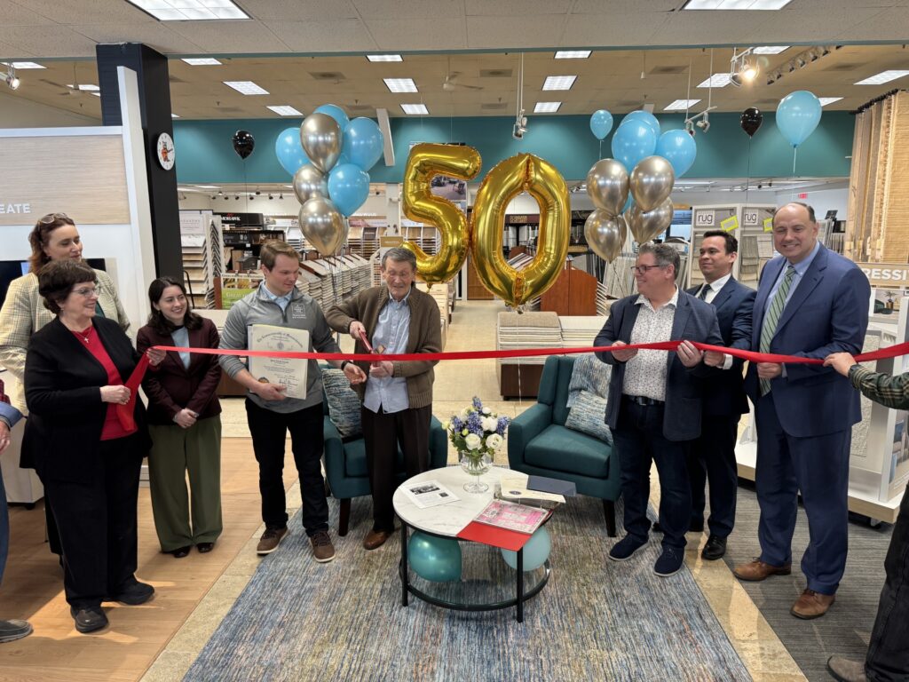 People holding a red ribbon while an older man wields oversized scissors. In the background, large gold balloons that say 50 are visible, along with many other blue and silver balloons.