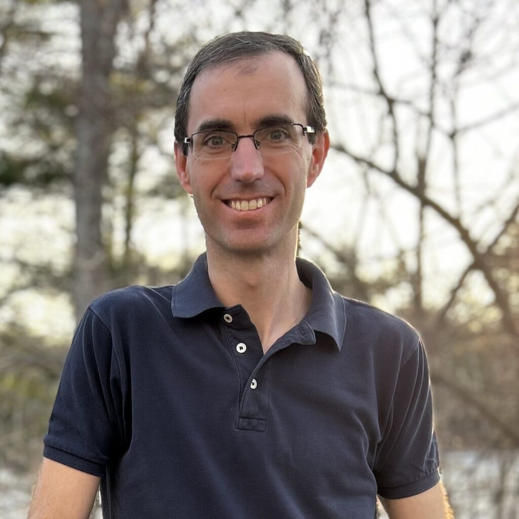 A smiling man wearing glasses stands outside with trees in the background.