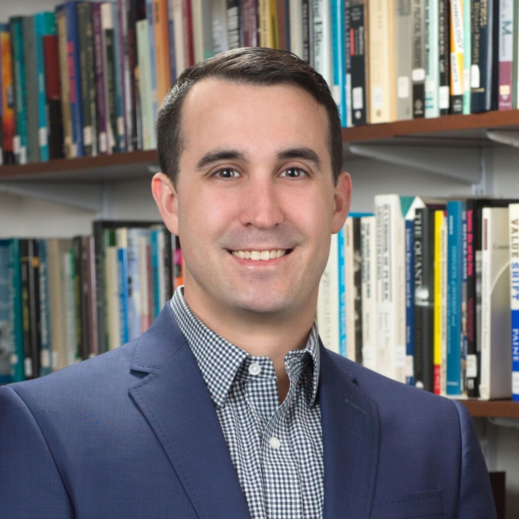 A man wearing a sportcoat and checked shirt stands in front of book stacks.