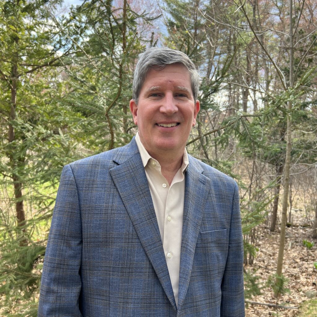 A man in a sport coat stands outside in front of some trees.