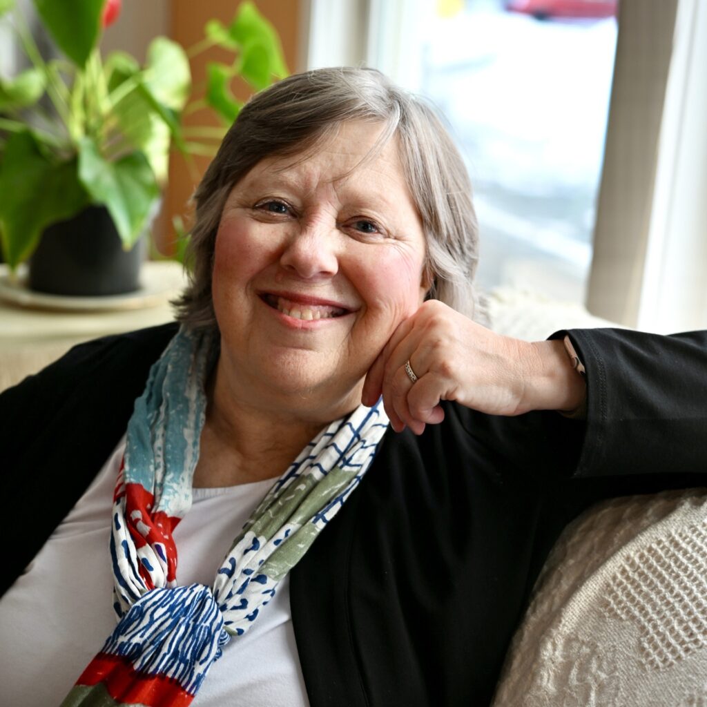 A relaxed-looking woman with short grey hair wears a balck sweater and colorful scarf around her neck.
