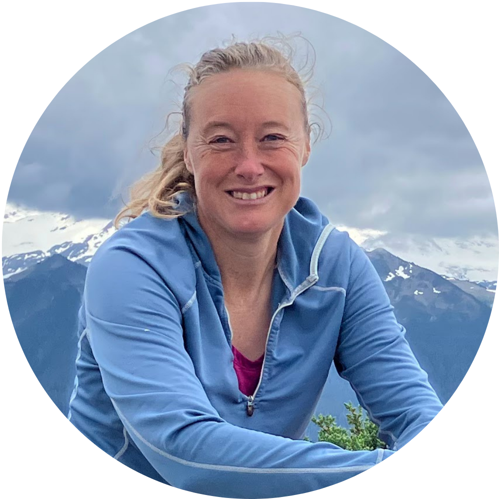 A smiling woman wearing a blue shirt with mountains in the background.