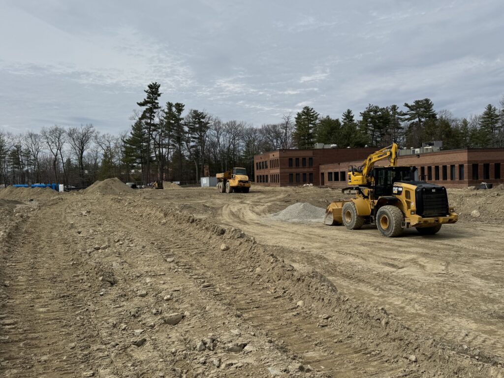 A brick building in the background with large construction equipment in front. Right now, it looks like the area is being flattened out before the next steps.