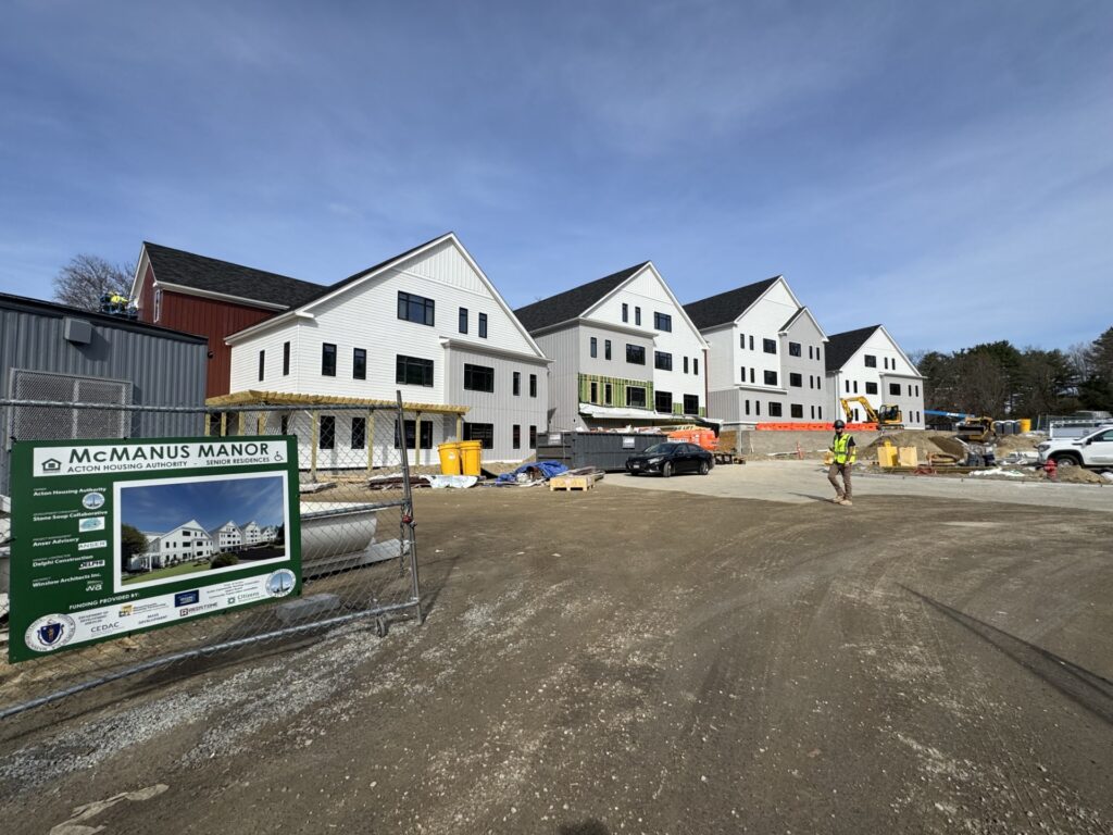 A housing development with four buildings is in progress. A man wearing a yellow vest and hard hat walks across what will be the parking area.