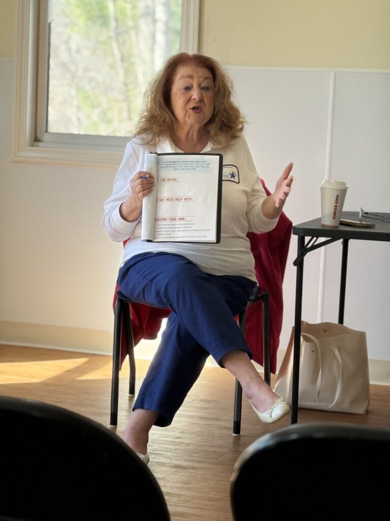 An older woman with shoulder length hair sits in a chair. She's holding a workbook turned to a page with places to enter information such as: "I can offer" and "I may need help with."