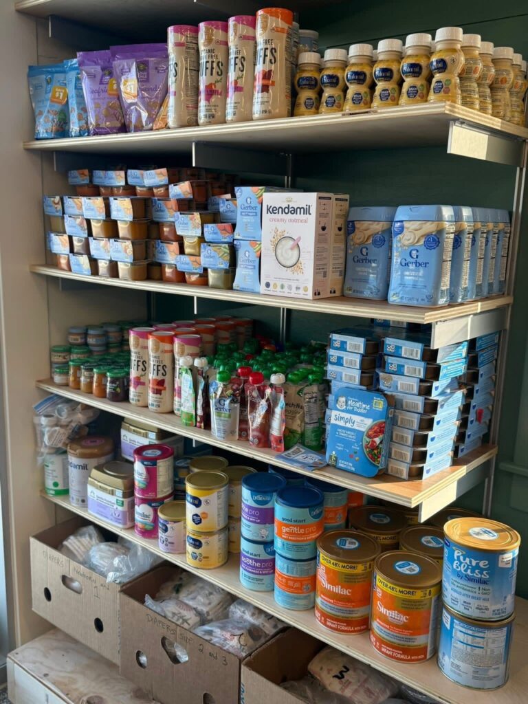 A shelving unit stocked with baby formula, baby food, and at the bottom, packets of diapers sorted by size.