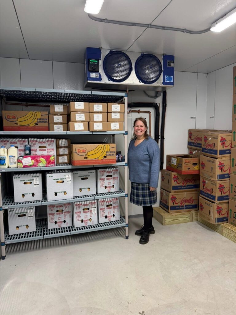 A woman wearing a blue sweater and plaid skirt stands next to a set of shelves that hold milk and produce. Abover her, a large cooling unit hangs frm the ceiling.