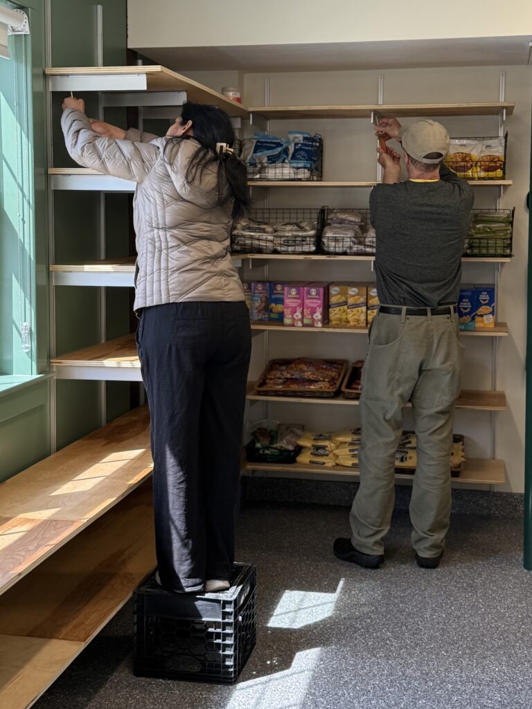 Two people screwing a high shelf into an existing set of shelves. One woman stands on a plastic crate to reach the shelf.