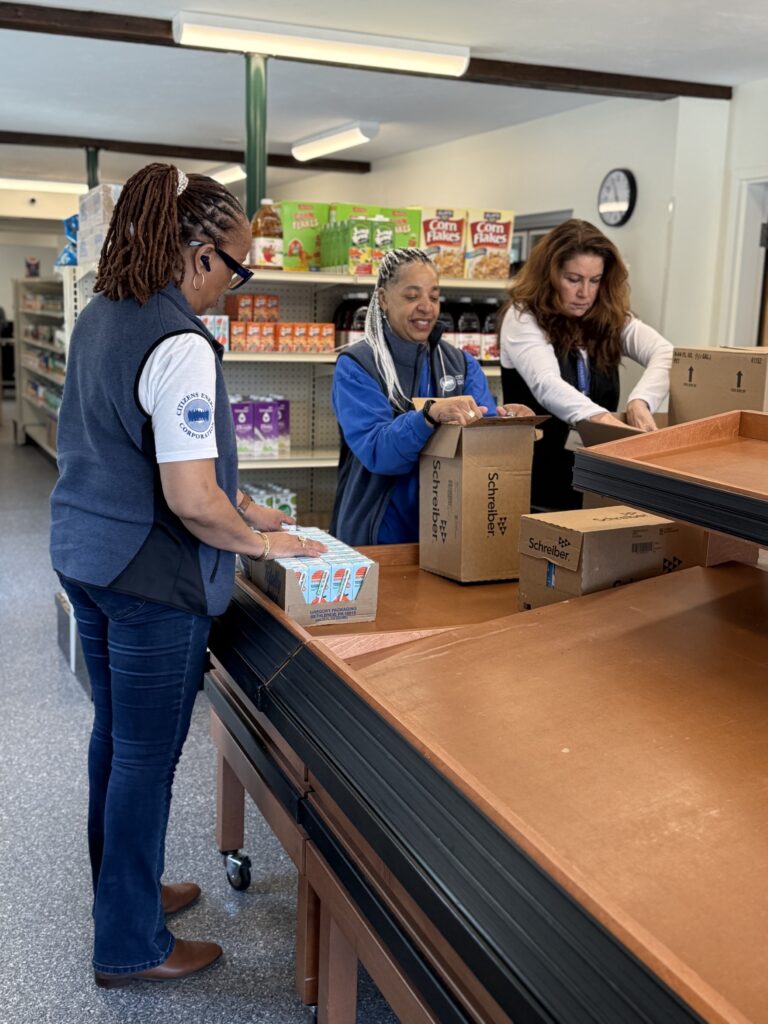 Three pepple stand at a table opening boxes of groceries. In the background are shelves with boxes of cereal and other staples.