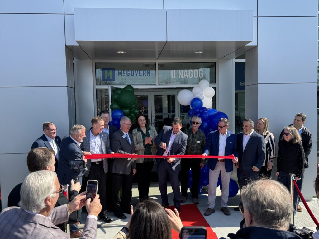 A group of people stand in front of the door holding a red ribbon. A man weilds the oversize scissors.