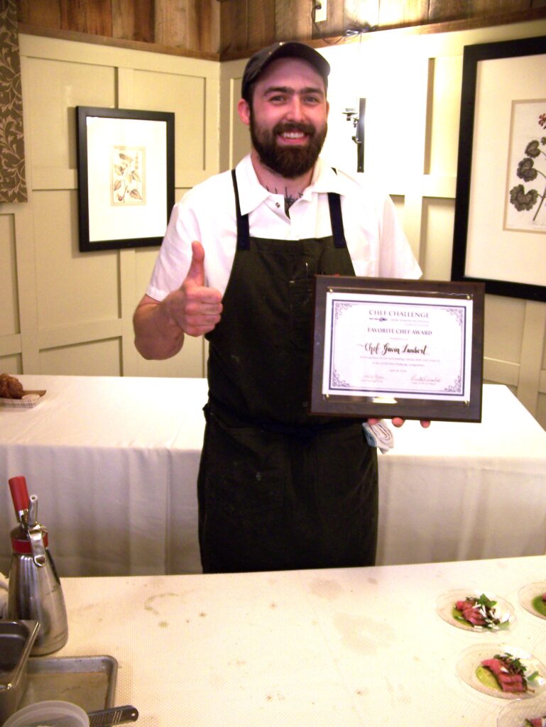 A man wearing an apron and ball cap stands in front of a white-clad dable. One hand is in a thumbs up sign, the other holds the Chef Challenge Favorite Chef Award.