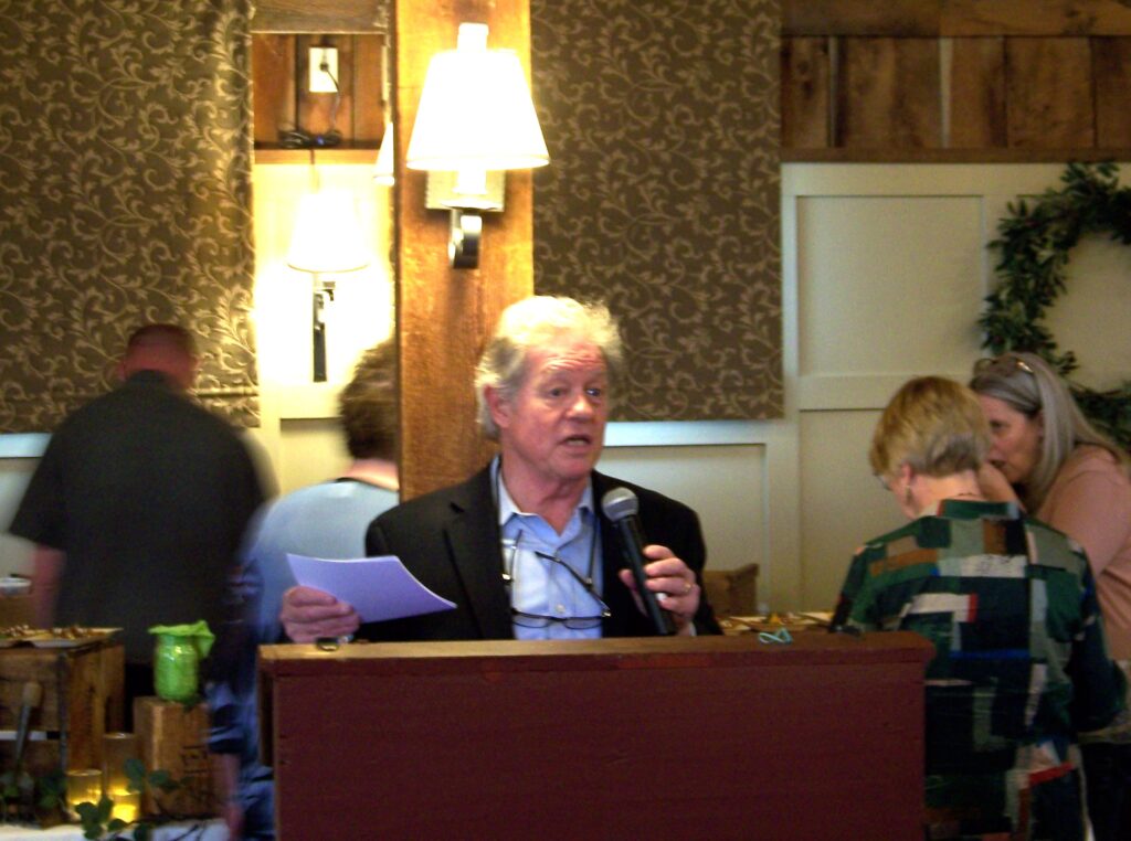 A man in a sport jacket speacks at a podium. A group of people sit at a table behind him.