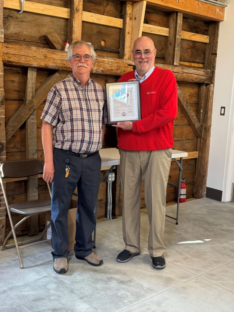 Two smiling men, one wearing a plaid buttondown shirt and the other in a bright red Rutgers sweater, hold a Faulkner Award plaque.