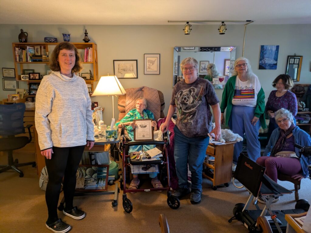 A tiny woman in a green shirt sits in a chair in a cozy room. She is surrounded by several other women.