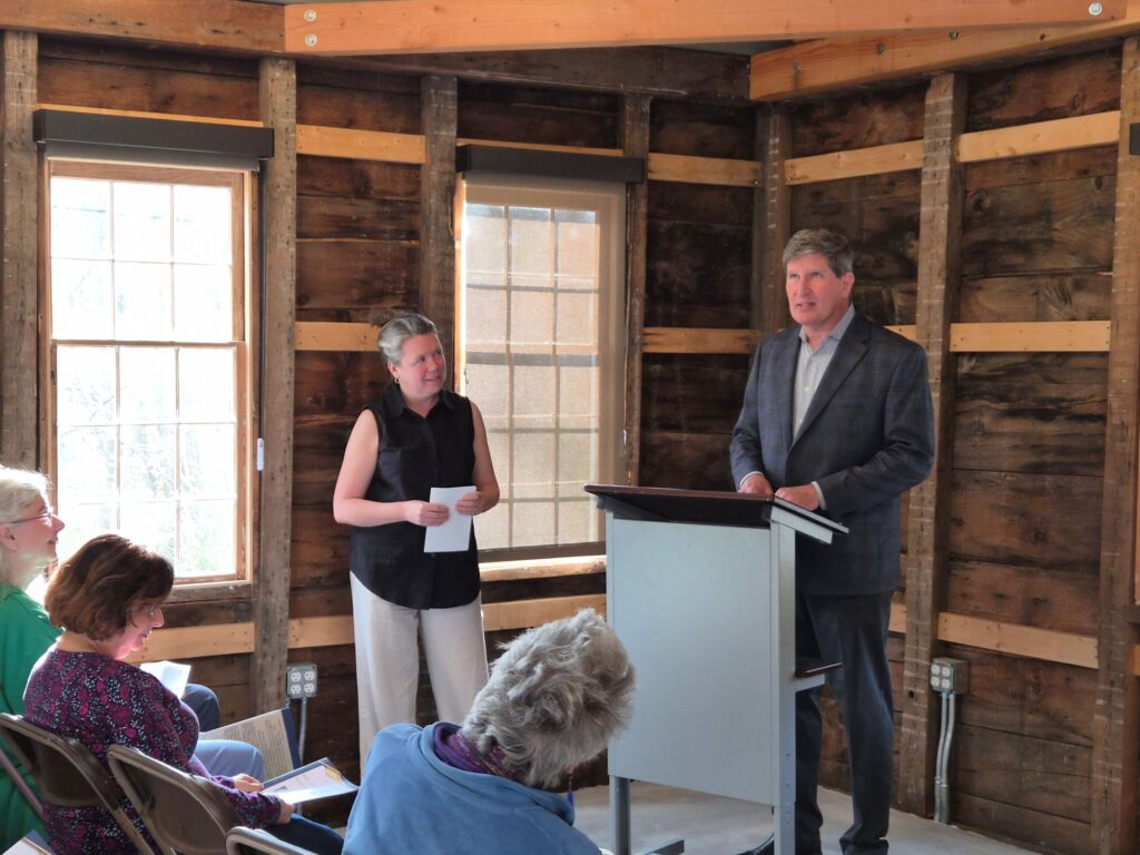 A man in a sport jacket speaks at teh podium while a woman in a black shirt looks on.
