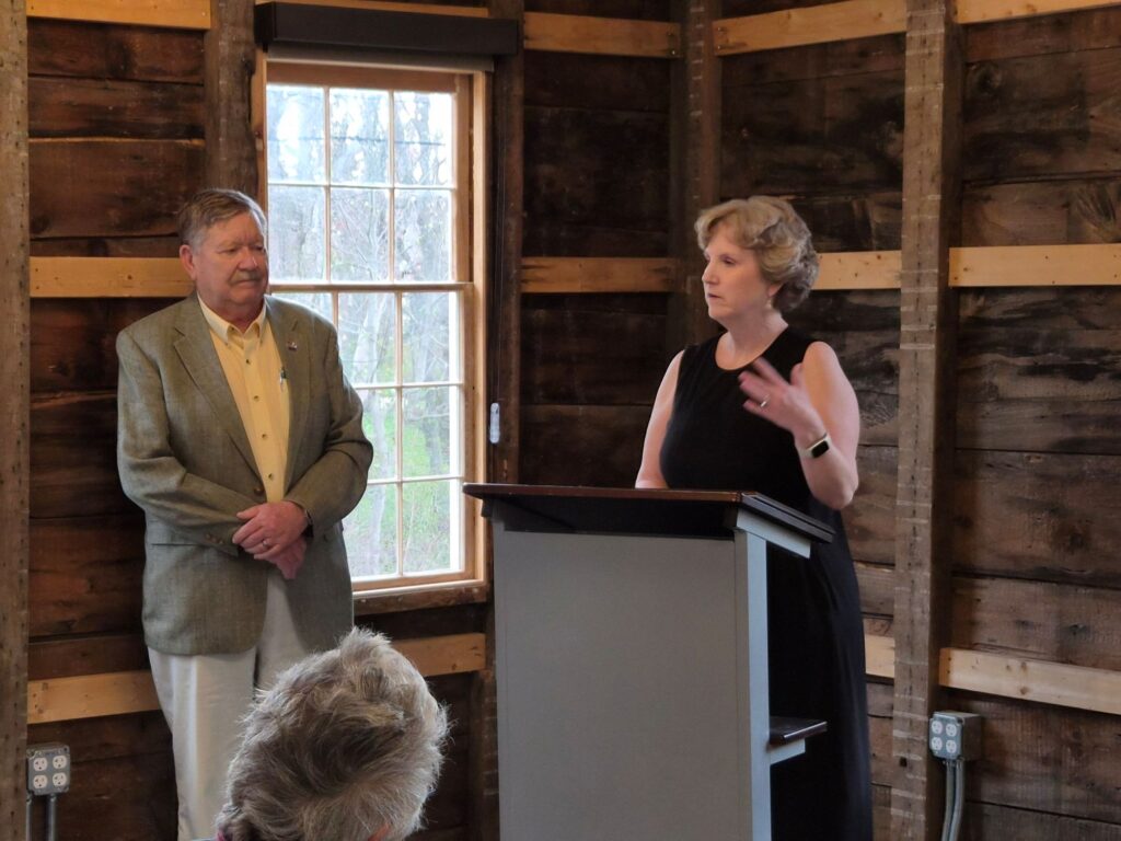A curly-haired woman in a black dress stands at a podium. A man in a sport jacket stands to her left, listening.