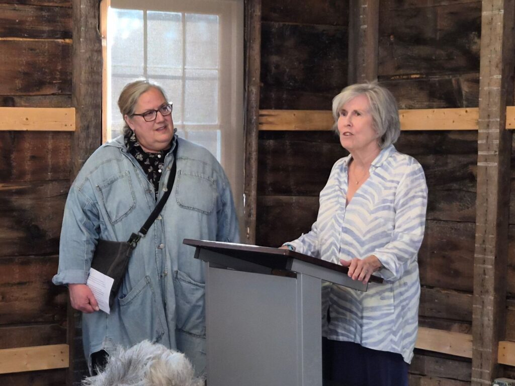 A woman with short grey hair speaks at the podium while anbother woman wearing a denim tunic looks on.