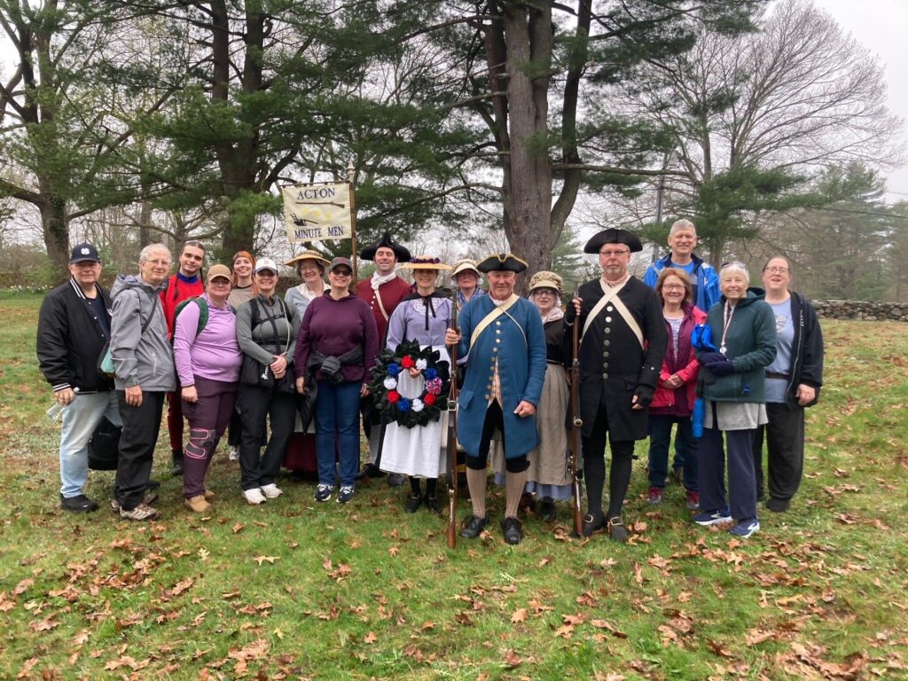 A group pf people stand under some trees. Some are in Colonial dress, some are not.
