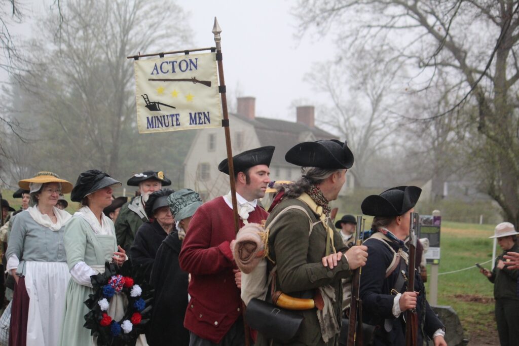 A group of people in Colonial garb are gathered, ready to march. One person carries a small Acton Minutemen flag that has a picture of a musket and Isaac Davis' plow.