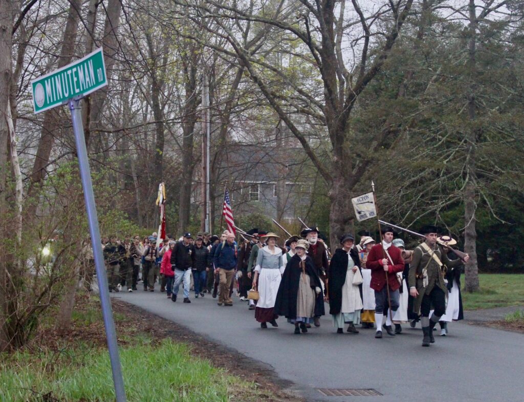 A group of people march down Minuteman Road in Acton. The men and women in front are in colonial garb, and some are carrying muskets.