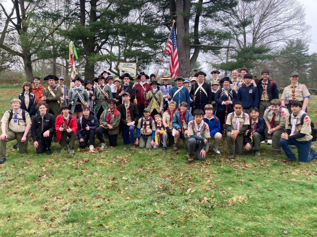 A large group of scouts pose with the Minutemen. The scouts carry an American flag and their troop flags; the Minutemen have their white flag.