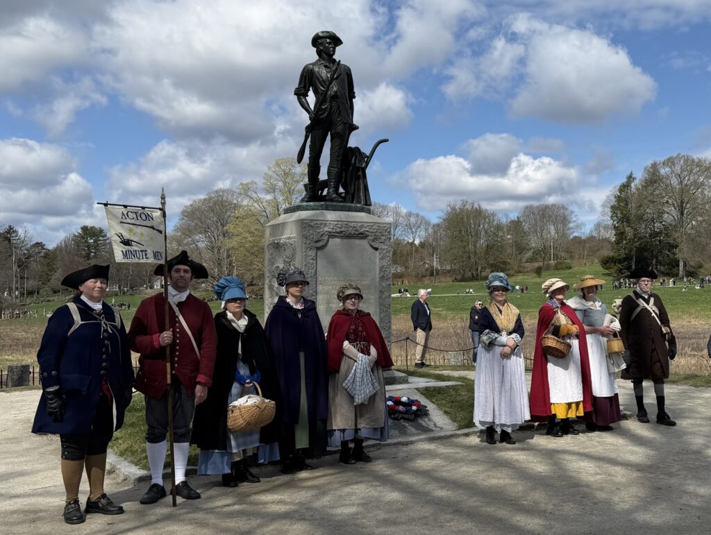 Some of the Acton Minutemen and women pose under a statue. The statue is on a stone pedestal, topped by a full-sized statue of a man carrying a musket and standing next to a plow.