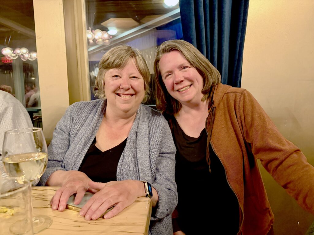 Two smiling women pose at a table. A glass of wine is in the foreground.