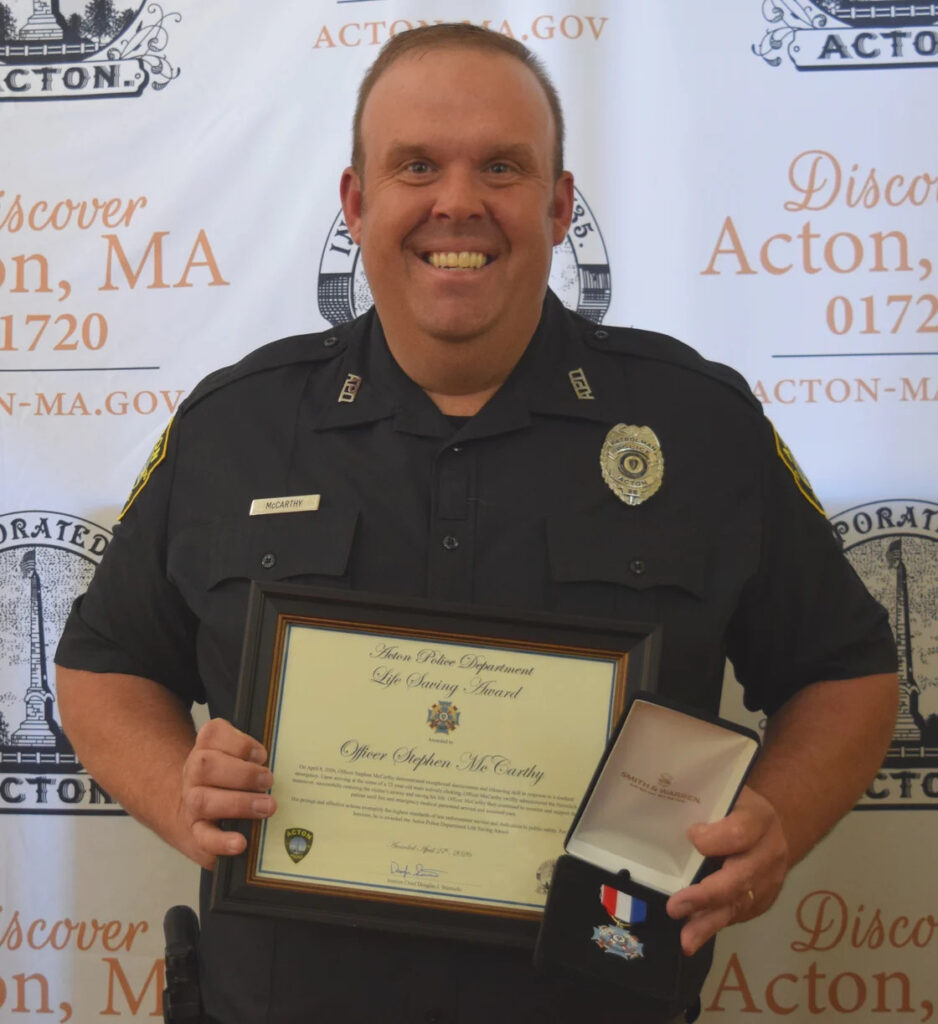 A smiling man in police unform holds a framed citatation and a medal while looking at the camera.